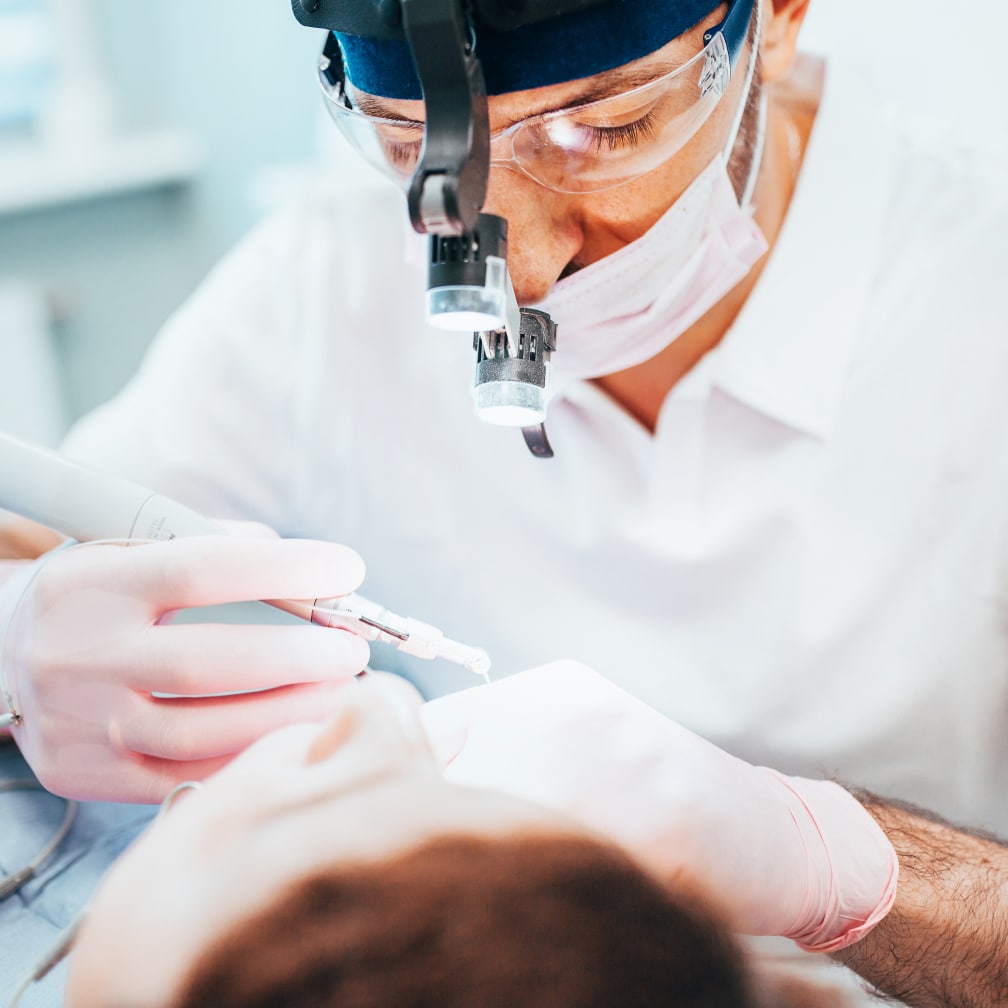 A person wearing a headlamp and protective gear performs dental work on a patient, focusing with precision instruments in a clinical setting.